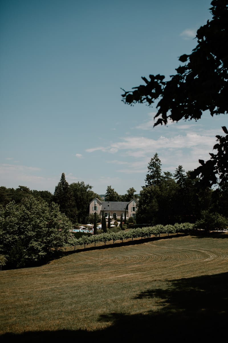 Wide landscape view of a French manor house surrounded by mature trees with white parasols on the terrace