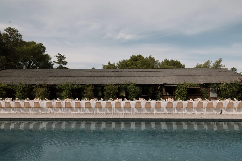 Long banquet table set on wooden pool deck with white linens, peach floral centerpieces, and thatched-roof venue behind