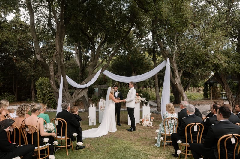 Outdoor ceremony under mature trees with white fabric canopy, couple at altar with candle columns and seated guests