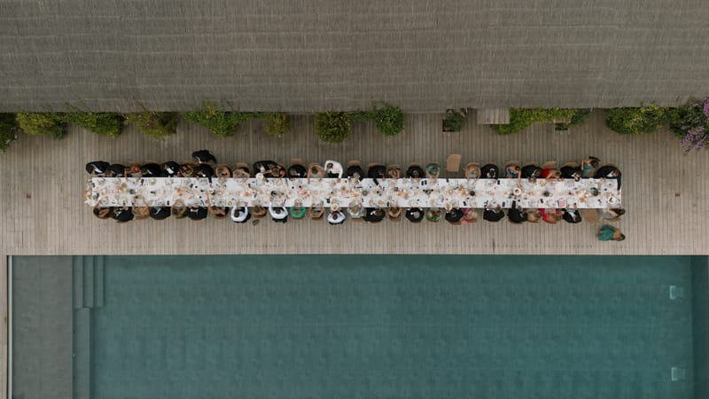 Aerial view of wedding reception dinner along a long table on wooden deck beside a swimming pool