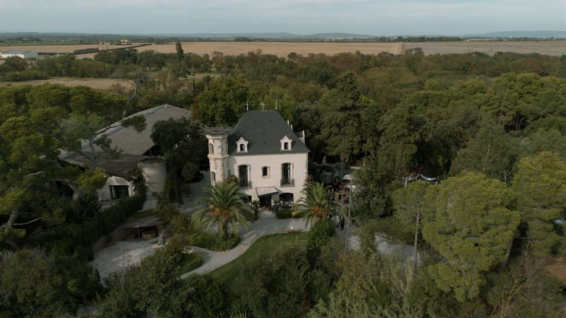 An aerial drone shot capturing a French country estate used as a wedding venue, surrounded by dense woodland with agricultural fields and low hills visible in the background. The main building is a white-rendered manor house with a dark grey mansard roof, dormer windows, and wrought iron balconies, consistent with late 19th-century French regional architecture. To the left, a large barn or reception structure with a corrugated metal roof is visible, along with what appears to be a circular stone tower or silo. A small group of wedding guests can be seen gathered to the right of the main building near a white marquee or canopy structure, suggesting a cocktail hour or outdoor reception in progress. Potential venue feature image.