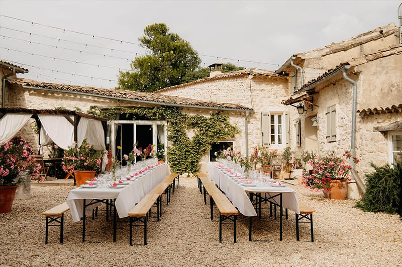 Outdoor reception with long tables and benches in gravel courtyard with festoon lights and stone buildings