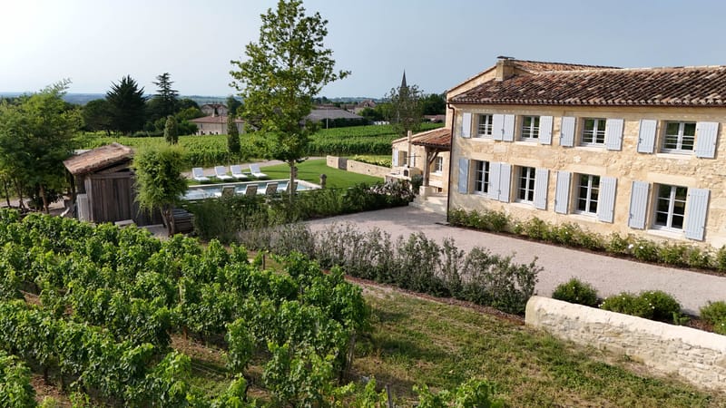 Aerial limestone property with blue-grey shutters, pool, and vineyard rows near village church spire