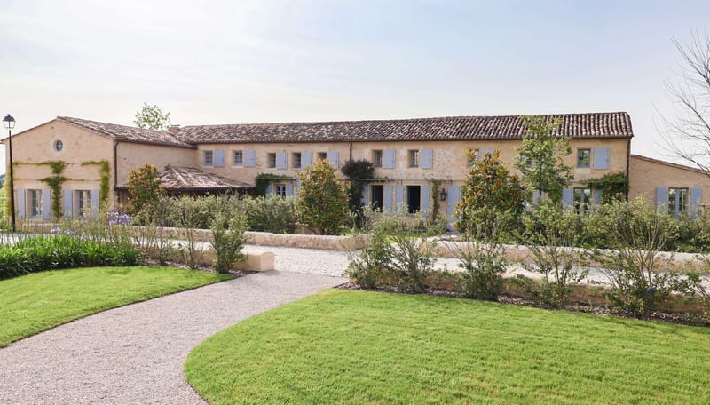 A wide exterior shot of a large French country estate or domaine, featuring a long two-storey golden limestone building with terracotta Roman tile roofing and pale blue-grey painted shutters across all windows. The facade is partially covered with climbing plants, and the entrance is framed by a wall-mounted lantern. The grounds in the foreground include a curved gravel driveway, low stone retaining walls, manicured lawn areas, young magnolia trees, ornamental shrubs, and purple flowering plants along the left border. No people are visible in the image. Potential venue feature image.