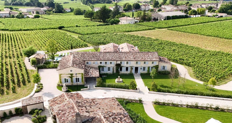 Aerial drone shot of a French country estate set within an expansive vineyard. The main building is a two-storey stone manor with terracotta tile roofing, pale blue shutters, climbing vines on the facade, and a central stone fountain in the gravel forecourt. The property includes multiple outbuildings with matching terracotta roofs, a swimming pool with sun loungers visible to the left, and well-maintained lawn areas bordered by low hedging and young trees. The surrounding landscape is entirely covered in neatly planted vineyard rows, with a village visible in the background. No people or wedding-specific decor are visible in this image. Potential venue feature image.