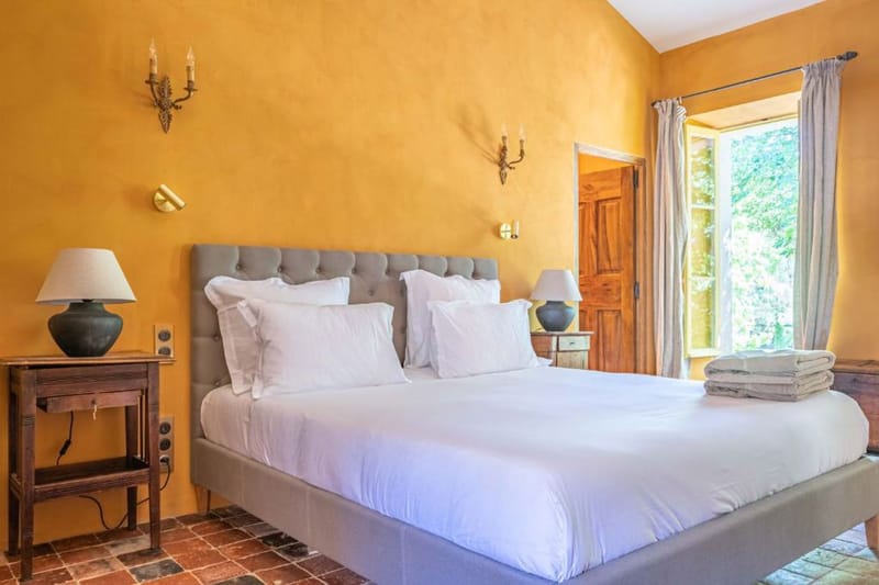 Provencal guest bedroom with ochre walls, grey tufted headboard, terracotta floors, and linen curtains