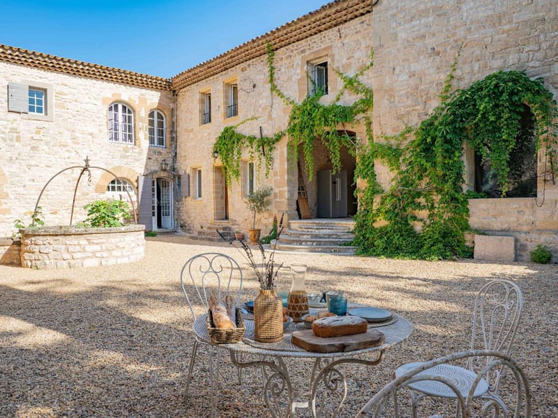 Provencal stone mas courtyard with arched doorways, climbing vines, stone well, and bistro table with French breakfast spread