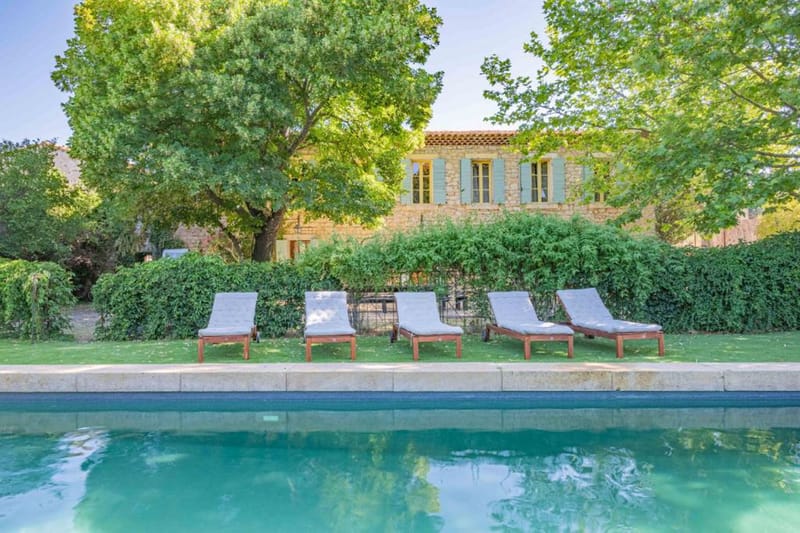 Wide exterior shot of a French stone mas or bastide-style property featuring a rectangular outdoor swimming pool in the foreground with turquoise water. Five wooden sun loungers with light grey cushions are lined up along the pool's stone coping edge. The two-storey stone building in the background has pale blue-green shutters and terracotta roof tiles, partially framed by mature trees. No people are visible in the image. Potential venue feature image.