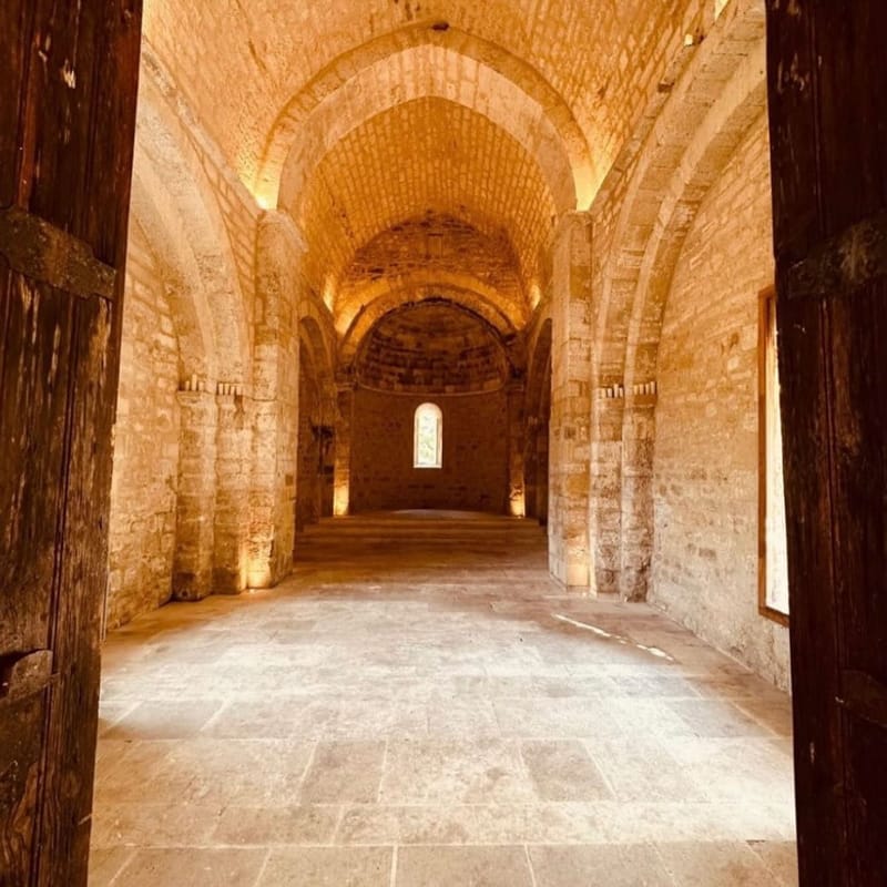 Interior shot of a Romanesque chapel or abbey taken from the entrance doorway, framed by large aged wooden doors. The space features barrel-vaulted stone ceilings, thick limestone columns with carved capitals, and a stone-flagged floor leading to an apse at the far end with a single arched window. Warm uplighting is placed at the base of the columns, casting a golden tone against the pale stone walls and vaulted ceiling. The room is currently empty with no wedding decor or guests visible. The composition is a central wide shot looking straight down the nave. Potential venue feature image.