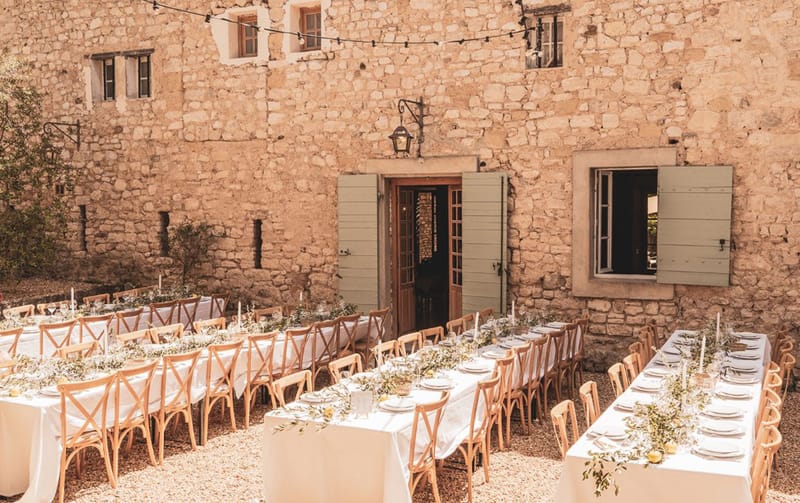 An outdoor wedding reception setup photographed in a wide shot, showing multiple long rectangular tables arranged in a courtyard in front of a large rustic stone building with sage green shutters and wooden doors. The tables are dressed in white linen with natural wood cross-back chairs on either side. Table runners consist of trailing olive branch greenery and what appear to be small yellow citrus fruits, with white taper candles and glassware placed throughout. A string of bistro lights is strung across the upper portion of the courtyard facade. The overall decor palette is white, natural wood, and muted green, consistent with a rustic Provençal style. No people are present — this is a pre-reception setup shot. Potential venue feature image.