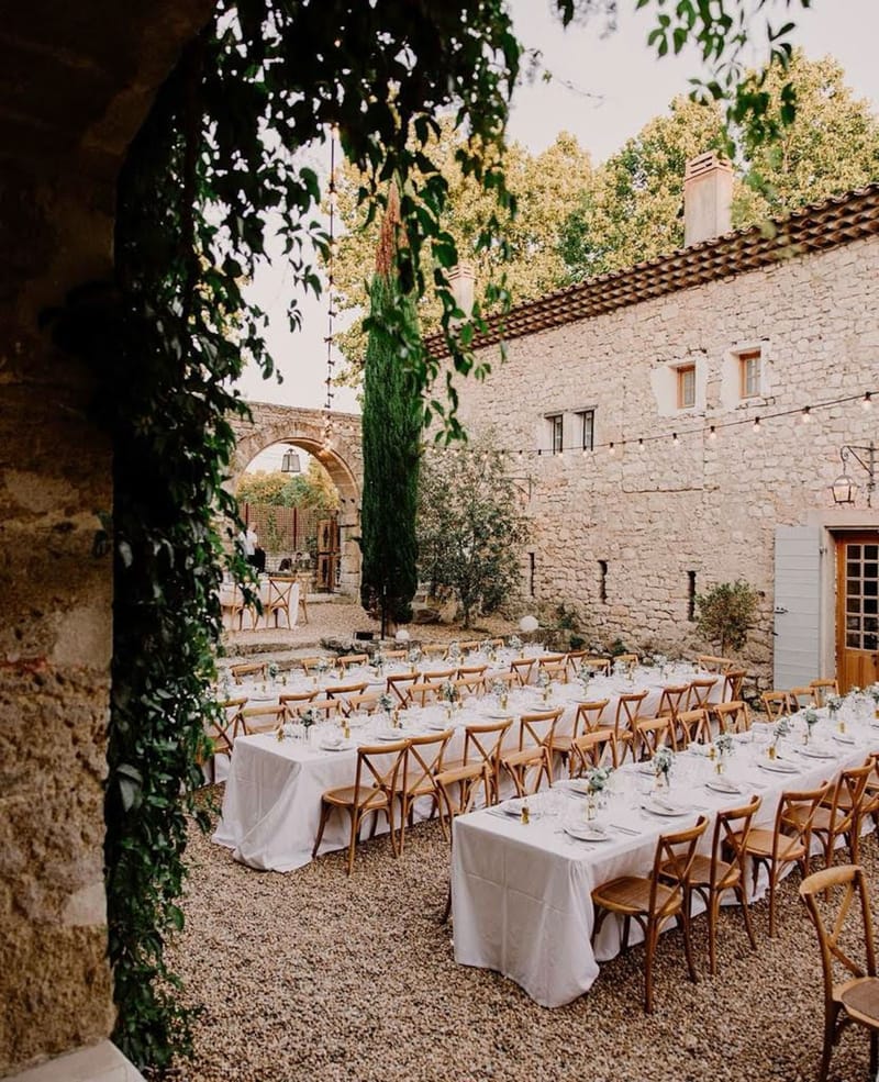 Outdoor reception setup in Provencal stone courtyard with long banquet tables, cross-back chairs, and festoon lights