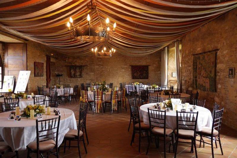 A wedding reception room set up for dinner inside a rustic stone-walled venue, likely a historic château or converted barn in France. The space features exposed stone walls hung with tapestries and framed artwork, terracotta tile flooring, and a wrought-iron chandelier with candle-style bulbs. The ceiling is draped with multicolored fabric panels in warm tones of orange, yellow, red, and gold, creating a tent-like canopy effect. Round tables are dressed in white linen and surrounded by dark wood chiavari chairs with taupe seat pads; centerpieces include yellow and green floral arrangements with pillar candles and small framed photos. A seating chart easel is visible near the entrance. Wide interior shot showing approximately eight to ten tables set for guests. Potential venue feature image.