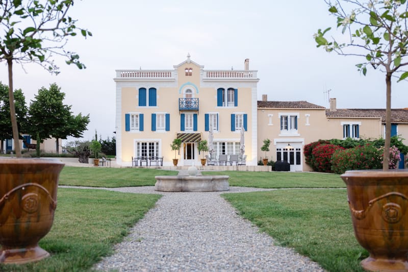 A wide exterior shot of a French country estate at dusk, with no wedding party or guests visible. The main building is a three-story pale yellow manor with blue shutters, a wrought-iron balcony on the second floor, and white-framed French doors on the ground level. A gravel pathway leads from the foreground directly to a stone fountain at the center of a manicured lawn, flanked by two large terracotta Anduze urns in the immediate foreground. Warm exterior lighting illuminates the facade of the manor. Adjacent buildings in the same pale yellow palette with matching blue shutters are visible to the right, bordered by red climbing roses. The composition is a symmetrical wide shot taken from ground level along the central axis of the property. Potential venue feature image.