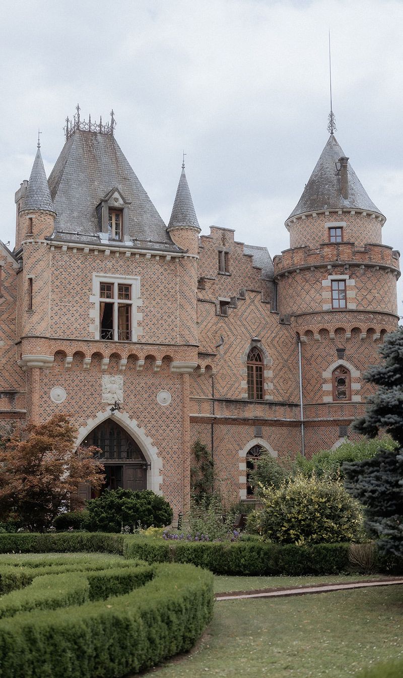 Exterior wide shot of a French château featuring warm terracotta and red brick construction with decorative diamond-pattern brickwork, multiple conical-roofed towers with grey slate finials, Gothic pointed archway entrance, and arched window details with white stone surrounds. The foreground includes formally clipped boxwood hedges arranged in geometric shapes, with ornamental shrubs and a blue-grey conifer to the right. No people are visible in the frame. The image is taken in natural overcast light. Potential venue feature image.