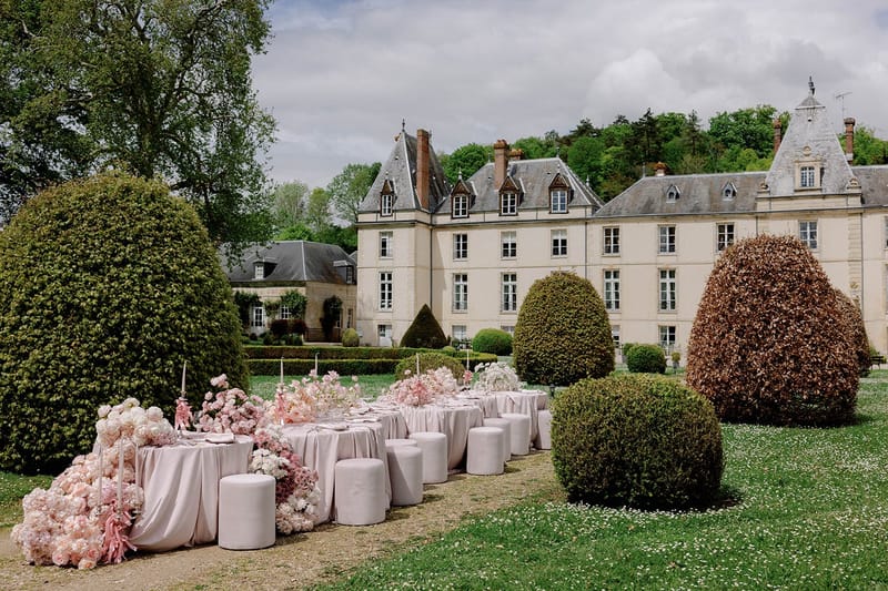 Blush-draped table with cascading pink peony arrangements and cylindrical ottoman seating before chateau