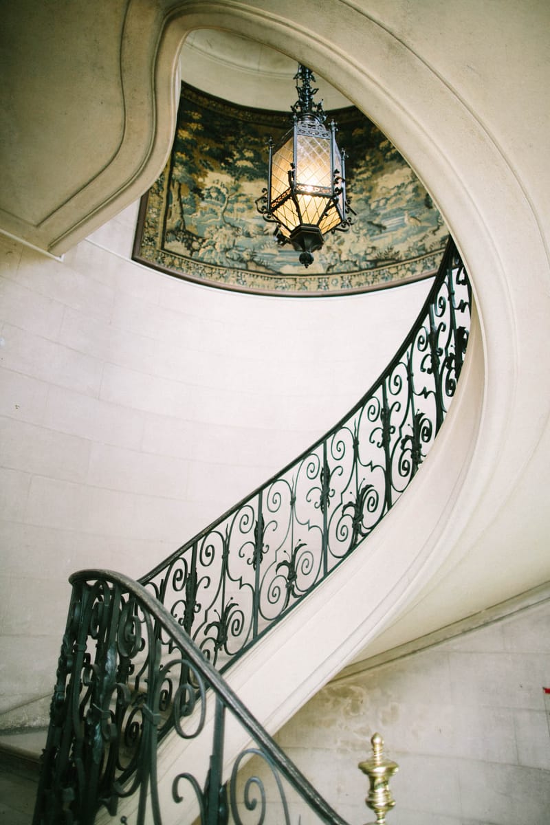 A detail shot of an indoor grand spiral staircase inside what appears to be a French château or historic manor. The staircase features an ornate dark wrought-iron balustrade with scrollwork detailing, set against cream-toned stone walls with curved classical molding. A decorative hanging lantern with amber glass panels is suspended from above, where a painted or tapestry-style ceiling mural depicting a pastoral scene in muted green and gold tones is visible through the oval stairwell opening. A small gold finial detail is visible at the lower right. No people are present in the frame. Potential venue feature image.