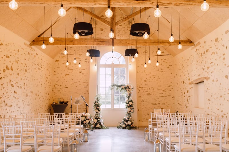 Indoor ceremony space with circular blush and ivory floral arch white Chiavari chairs and Edison pendant lights