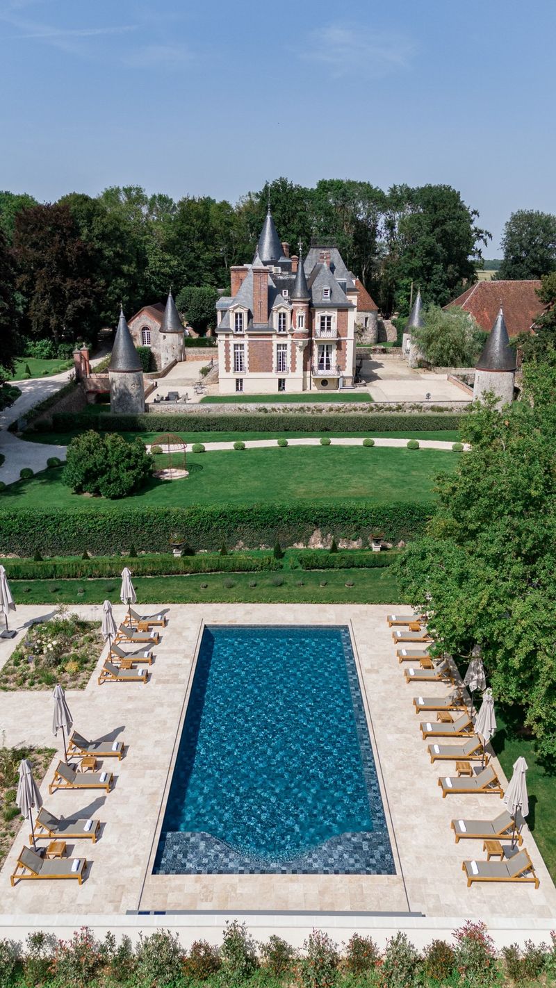 Aerial multi-turreted brick chateau with formal box garden swimming pool limestone terrace and teak loungers
