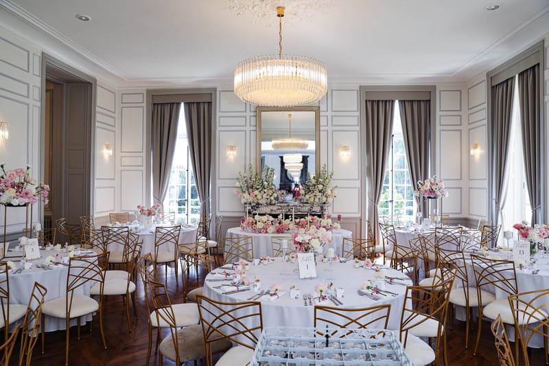 Chateau ballroom with crystal chandelier round tables gold chairs and blush pink floral arrangements