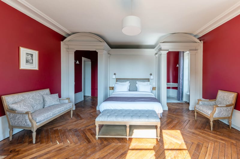 Crimson-walled chateau bedroom suite with arched doorways, herringbone parquet, and grey tufted bench