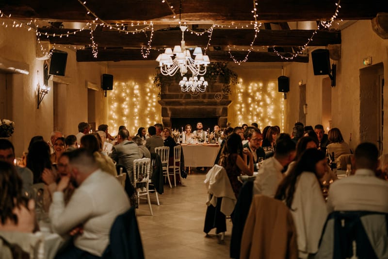 A wedding reception dinner is underway inside a rustic indoor hall with exposed wooden ceiling beams and stone walls. The couple is seated at a sweetheart table centered at the far end of the room, positioned in front of a large stone fireplace flanked by dense curtains of warm white fairy lights that cover the back wall. Two white chandelier-style pendant lights hang from the ceiling, and additional strings of fairy lights are draped across the wooden beams overhead, creating a warm amber glow throughout the space. Approximately 60–80 guests are seated at long rectangular tables arranged in two parallel rows along either side of a central aisle, using white Chiavari chairs, with candles and floral centerpieces visible on the tables. The overall styling is rustic with warm, candlelit tones, and the wide-shot composition captures the full length of the room from the entrance toward the couple's table.