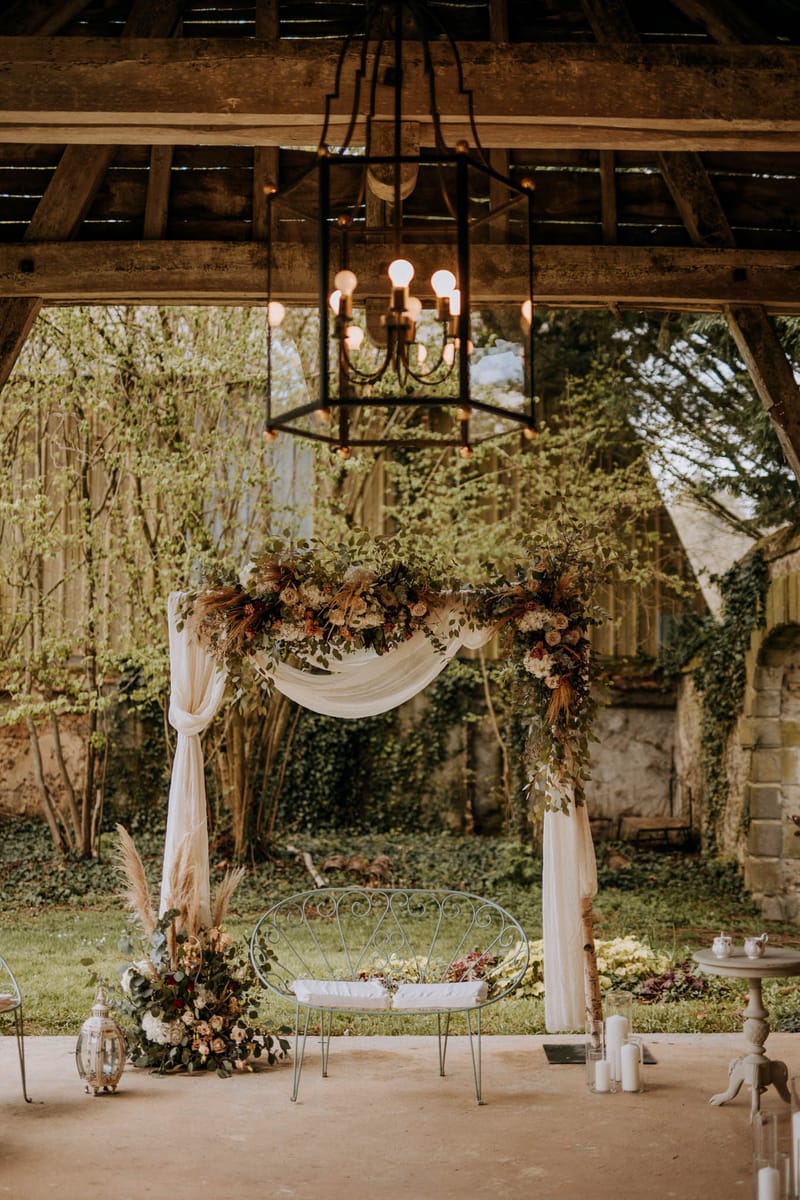 A ceremony setup photographed in wide shot beneath a rustic open-sided structure with exposed wooden beam rafters. A large black iron lantern chandelier with Edison-style bulbs hangs centrally above the altar area. The ceremony arch is decorated with lush greenery, ivory hydrangeas, blush and terracotta roses, burgundy blooms, and dried pampas grass, with ivory fabric draping loosely across its width. A vintage-style white scrollwork iron loveseat with a white cushion is positioned in front of the arch as the couple's seat. Ground-level floral arrangements at the base of the arch echo the same color palette of blush, terracotta, burgundy, and ivory with eucalyptus. To the left, a glass lantern with white pillar candles sits on the floor; to the right, a small antique side table holds what appears to be a tea service and white pillar candles. The overall styling is rustic-boho with warm, earthy tones. Potential venue feature image.