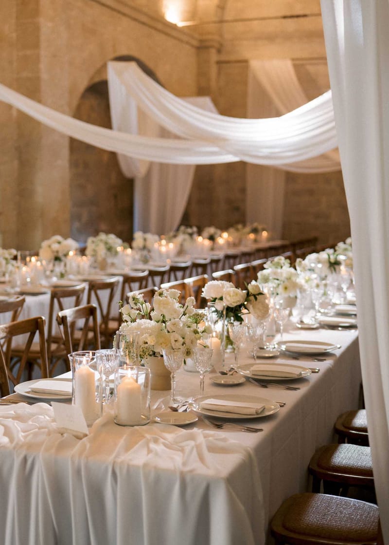 Stone-walled reception with sheer ceiling drapes white peony centerpieces cross-back chairs and pillar candles