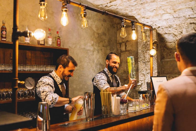 A cocktail hour scene set in an indoor cave-like bar with exposed rough stone walls and vaulted ceiling. Two bartenders dressed in matching dark waistcoats over floral-print shirts and patterned ties are actively preparing cocktails behind a dark wood bar counter; one is shaking a mixer holding a pale yellow drink, while the other smiles and stirs a tall green cocktail. The bar is equipped with a wood shelving unit stocked with spirit bottles including Havana Club rum and Stolichnaya vodka, rows of coupe and martini glasses, and a collection of mixing tools, straws, and garnish cups arranged on the counter. Lighting is provided by exposed Edison bulb pendants mounted on copper pipe rigs, casting a warm amber glow throughout the space. A guest in a blush-toned jacket is visible from behind in the foreground, and a small framed 'Cocktails' sign is displayed on the bar. The overall styling is industrial-rustic. Medium shot with slight bokeh on the foreground guest.