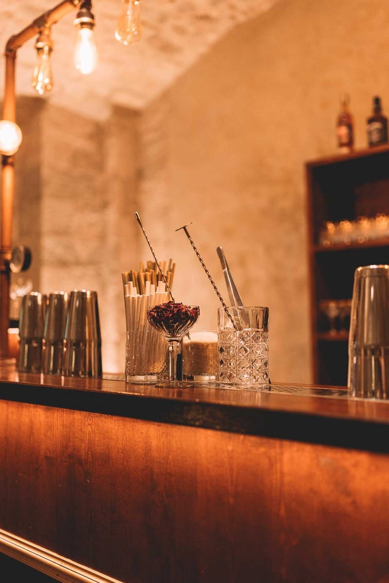 Close-up detail shot of a bar setup during what appears to be a wedding cocktail hour or reception, set in an indoor venue with exposed stone walls. The warm-toned wooden bar counter holds crystal cut-glass tumblers, metal cocktail shakers, bar spoons with twisted handles, paper straws in a tall glass, and a small coupe glass filled with dried dark red flower petals used as a garnish station. Overhead, bare Edison-style bulbs hang from copper pipe fixtures, casting warm amber light across the scene. In the background, a dark wood shelving unit displays spirit bottles including recognizable whisky labels, alongside glassware. The overall styling is industrial-rustic with a warm, candlelit palette of copper, amber, and gold tones.