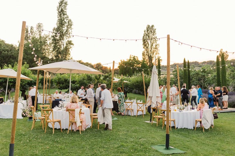 An outdoor wedding reception dinner is underway on a garden lawn at golden hour, with approximately 30–40 guests mingling and seated across multiple round tables. The tables are dressed in white linen and surrounded by natural wood cross-back chairs, with low floral centerpieces featuring blush pink and white blooms, candles, and glassware. Overhead, bistro globe string lights are strung between tall wooden poles, and large cream patio umbrellas provide shade over several tables. A small wooden dance floor is visible in the background on the right. The styling is relaxed and rustic-garden in tone, with a warm neutral palette of white, natural wood, and soft blush. Guests are dressed in a mix of smart-casual and formal attire, including floral print dresses, a blue dress, and a black tuxedo. The wide-angle shot captures the full reception layout from a slight elevation, conveying the overall scale and atmosphere of the setting.