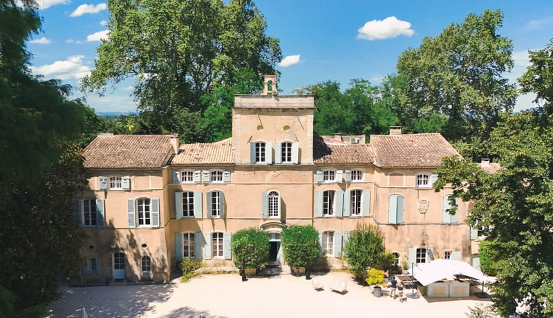 An aerial wide shot of a French château with warm terracotta-pink rendered façade, terracotta tile roof, and pale blue-grey shutters across multiple windows. The three-storey building features a central tower element with a sundial, arched entrance doorway framed by trimmed green hedging, and a stone coat-of-arms detail on the right wing. The gravelled forecourt in front of the building includes potted plants, a few white outdoor chairs, and a white market umbrella with two people visible nearby. The property is surrounded by mature deciduous trees on all sides. Potential venue feature image.