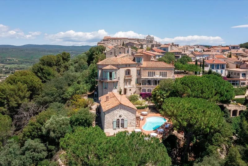 Aerial drone shot of a historic stone property with terracotta-tiled roofs, teal-painted shutters, and an outdoor pool with white parasols and seating visible in the lower section of the grounds. The main building is a multi-story Provençal-style stone manor surrounded by mature trees, with a smaller stone chapel-like structure in the foreground. The property sits within a hilltop village featuring densely packed stone and rendered buildings with matching terracotta rooftops, a church visible at the summit, and an open valley with agricultural land stretching into the distance. Potential venue feature image.