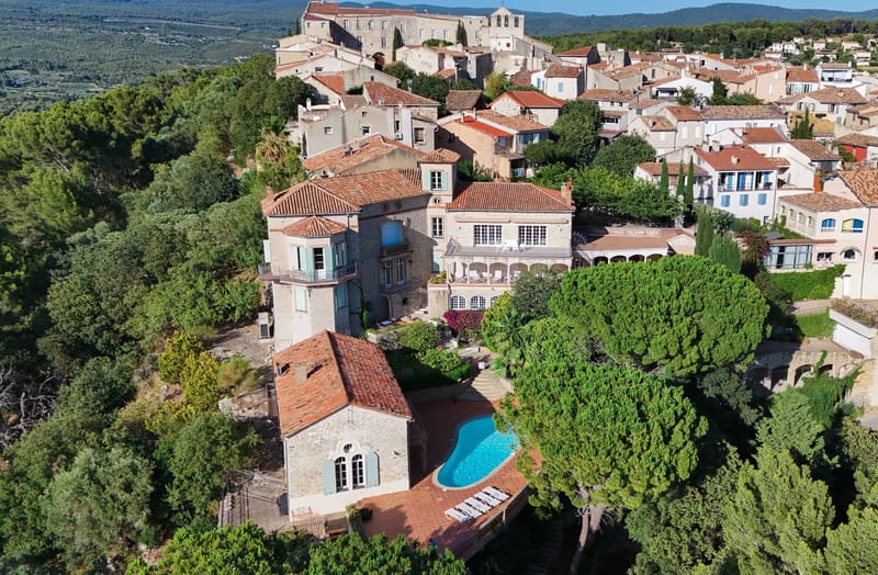 Aerial drone shot of a stone manor house with pale blue-green shutters situated within a densely built Provençal hilltop village, with terracotta-tiled rooftops extending across the hillside and a large historic church or château visible at the summit. The property features a freeform swimming pool with a terracotta tile surround and white sun loungers, a terraced courtyard with visible pink-flowering bougainvillea, and a multi-building stone complex with arched colonnades on the main façade. The surrounding village shows uniform terracotta roof tiles and pale stone construction typical of southern France, with forested hillsides in the background. No people are visible in the image. Potential venue feature image.