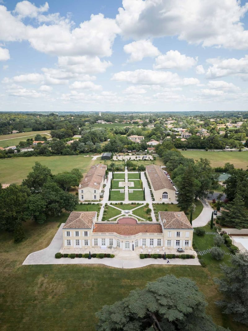 Aerial drone view of French chateau with terracotta roof, formal parterre garden, cypress trees, and outbuildings