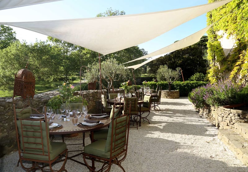 An outdoor reception dining setup on a gravel terrace, shaded by large white triangular sail canopies stretched overhead. Multiple round iron tables are set for dining with wine glasses, white folded napkins on woven charger plates, and small floral centerpieces with pink blooms. The wrought-iron chairs have sage green cushioned seats and backs. Decorative rust-patinated Moroccan-style lanterns are placed on the tables and mounted on curved iron stands throughout the space. The terrace is bordered by low dry-stone walls with lavender plantings and trimmed hedges along the right side. No people are present in the image. The overall styling is Provençal with a relaxed, rustic-Mediterranean aesthetic. Wide shot taken from a low angle looking down the length of the terrace. Potential venue feature image.