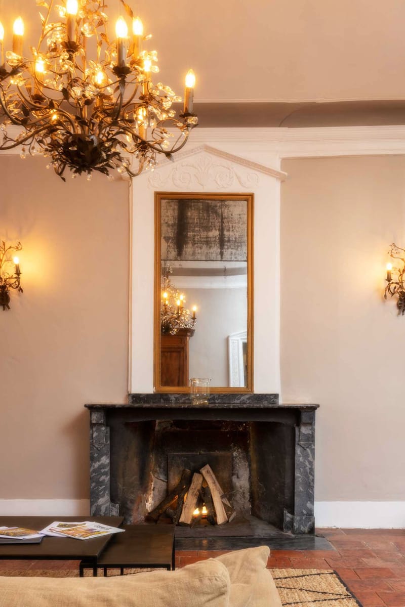 Interior shot of a château reception room featuring a dark grey marble fireplace with logs set inside, topped by a tall gold-framed antique mirror set within an ornate white plasterwork alcove with carved decorative detailing. The room is lit by a wrought-iron branch-style chandelier with crystal accents and candle-style bulbs in the upper left, and matching wall sconces on either side of the fireplace. The walls are painted in a warm off-white or pale blush tone, and the floor is terracotta tile partially covered by a natural woven rug. A low black coffee table with magazines and a cream upholstered seat are visible in the foreground. No people are present. Potential venue feature image.