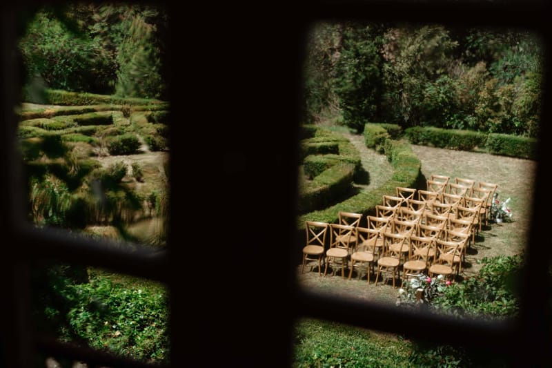 Aerial view through window of outdoor ceremony setup with cross-back chairs and boxwood hedges in French garden