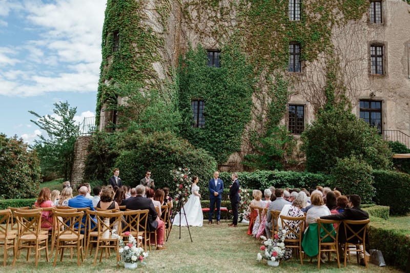 60 guests at ceremony before ivy-covered chateau with red and white floral arch and aisle markers