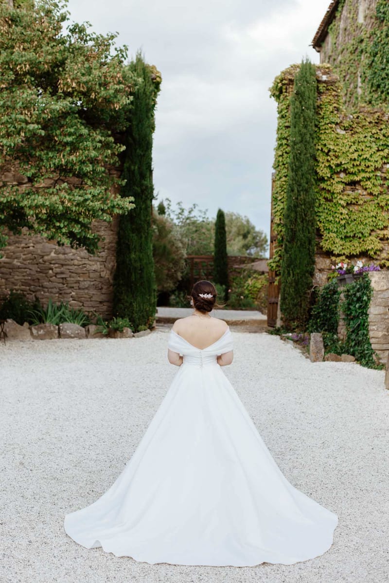 Bride from behind in off-shoulder ballgown with cathedral train spread on gravel courtyard flanked by cypress trees