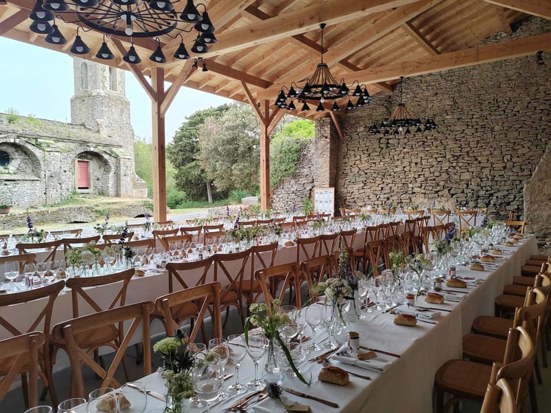 Long banquet tables set under a wooden-beamed pavilion with wildflower centerpieces and black pendant chandeliers