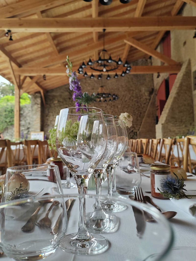 A close-up detail shot of a wedding reception table setting inside a rustic barn or covered open-sided structure with exposed natural wood beam ceiling and stone walls. The foreground shows clear wine glasses, bronze/copper-toned cutlery, white linen tablecloths, small glass jars of what appears to be honey or jam with gold lids used as favors, and blue thistle and cream scabiosa floral accents as individual place settings. In the background, loose wildflower centerpieces include purple delphiniums, green foliage, and white blooms in small vases. Natural wood cross-back chairs line the tables. Two black wrought-iron circular chandeliers hang from the wooden roof structure, and the overall decor palette follows a rustic-botanical theme with wildflower arrangements, natural materials, and warm tones.