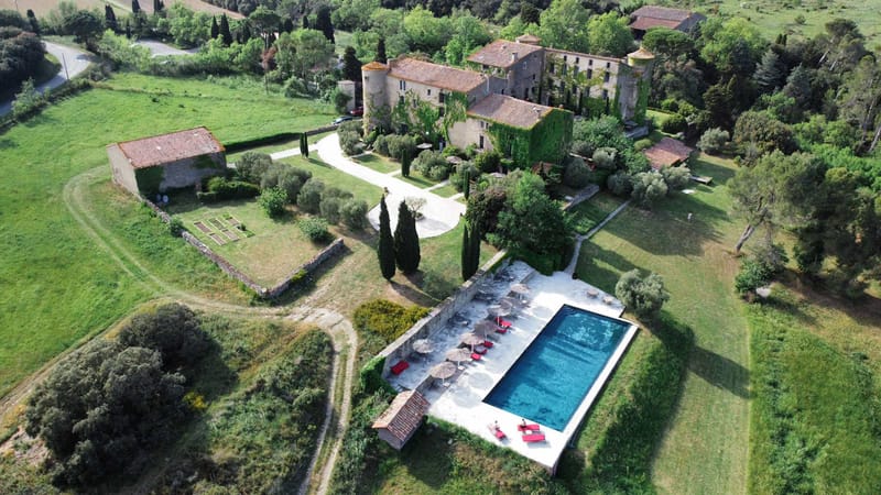 Aerial view of ivy-covered stone chateau with swimming pool, cypress-lined driveway, and surrounding fields