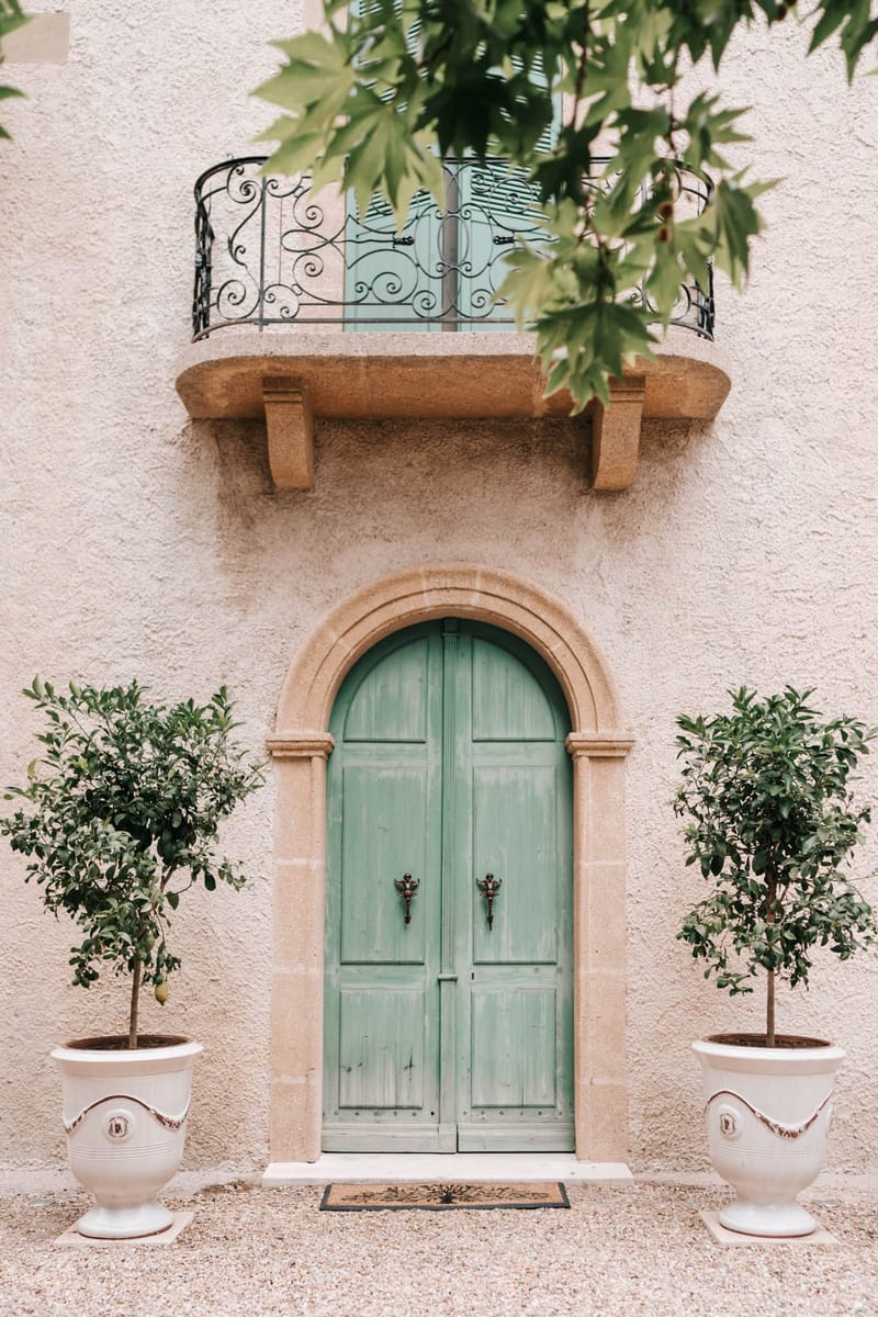 Provencal chateau entrance with sage green arched doors, limestone archway, wrought-iron balcony, and topiary citrus trees