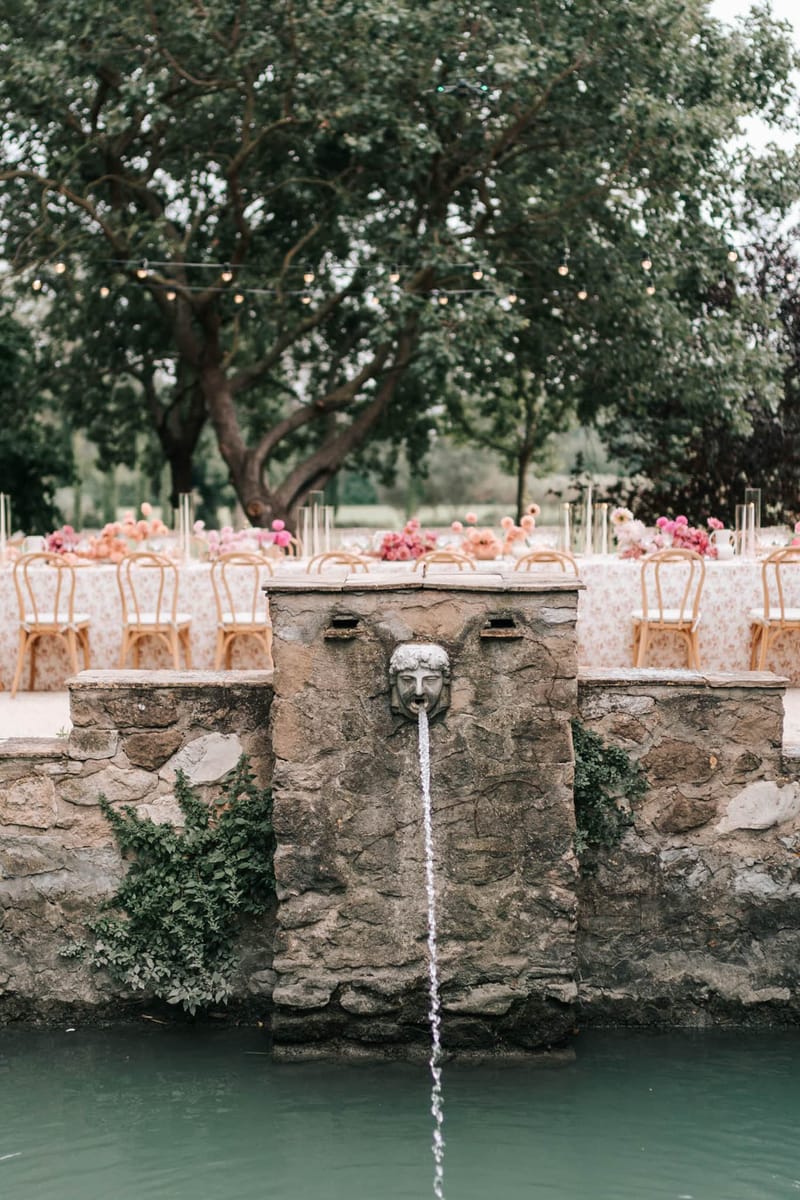 Outdoor reception table with pink floral tablecloth and peony arrangements viewed across pool fountain