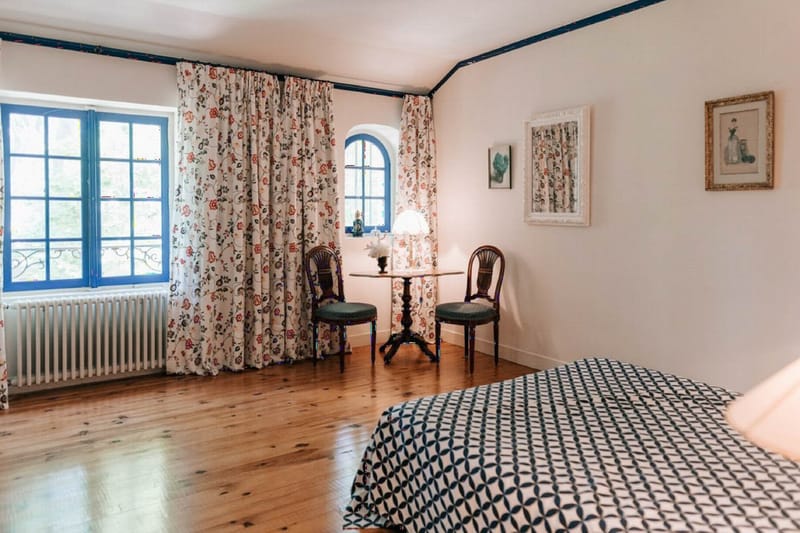 Chateau bedroom with honey hardwood floors, navy blue trim, arched window, floral curtains, and French country furnishings