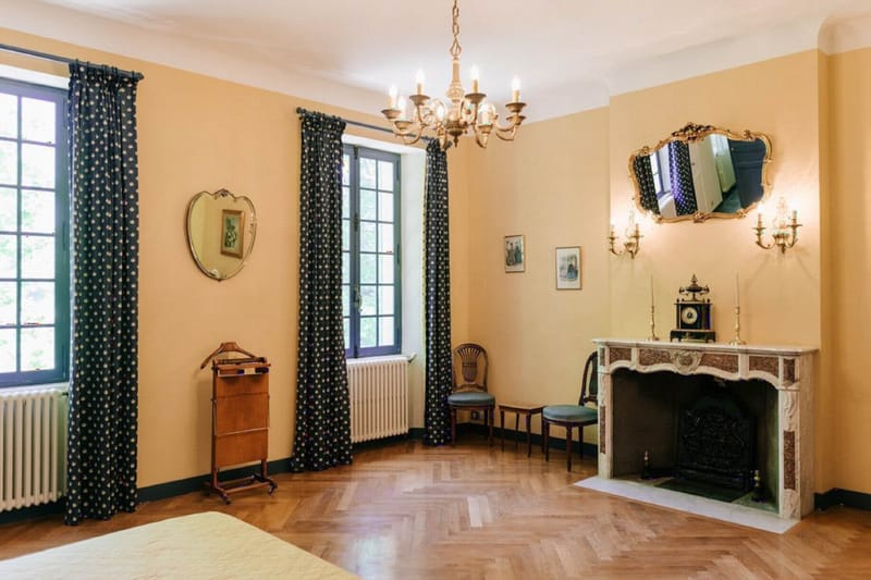 Interior wide shot of a bedroom inside what appears to be a French château or manor house, showing no people or wedding activity. The room features warm ochre-yellow painted walls, herringbone parquet flooring, and a white marble fireplace with carved detailing and a black iron insert, topped with a mantel clock, brass candlesticks, and wall-mounted gilt sconces. Dark navy curtains with a small white dot pattern hang at two large multi-pane windows. Furnishings include two upholstered chairs in sage green fabric, a wooden valet stand, and a partial view of a bed with a pale yellow cover. A brass chandelier with candle-style lights hangs from the ceiling, and two mirrors — one heart-shaped and one ornate gilt-framed — are mounted on the walls alongside small framed prints. The overall interior style is classic French period décor. Potential venue feature image.