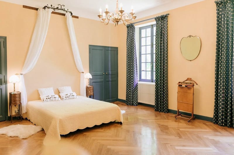Interior wide shot of a château bedroom, likely used as a bridal suite or getting-ready space. The room features warm peach-painted walls, herringbone parquet flooring, and a double bed dressed in a cream quilted coverlet with white embroidered pillows. A decorative canopy with white draping panels is mounted above the bed using a wrought-iron bracket. The room is lit by a brass multi-arm chandelier and two bedside table lamps. Dark teal-green painted doors, baseboards, and polka-dot curtains provide a contrasting color accent throughout the space. Furnishings include two small antique wooden nightstands, a wooden valet stand, and a shield-shaped wall mirror. A tall French window with blue-grey painted trim allows natural light into the room. The overall styling is classic French country with vintage furnishings. Potential venue feature image.