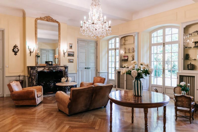 A wide interior shot of a classic French château salon with no people present. The room features a crystal chandelier, a black marble fireplace with a large gold-framed mirror above it, pale yellow walls with white panelled doors, and herringbone parquet flooring. Tan leather club chairs and a sofa are arranged around a small marble-topped table near the fireplace, while a round polished wood dining table with cane-backed chairs sits in the foreground. A striped green and white ceramic vase holding white peonies or ranunculus is placed on the round table, with a smaller white floral arrangement visible in the background. Natural light enters through tall arched French doors with a view of greenery outside. The decor style is classic French provincial with antique furnishings and ornate wall sconces. Potential venue feature image.