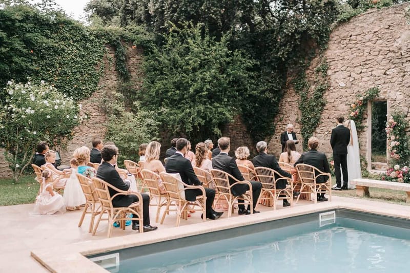 30 guests in rattan chairs at poolside ceremony with peach and white floral arch beside stone building