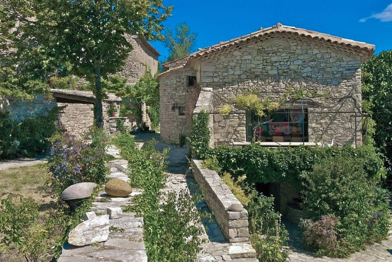 An outdoor wide shot of a traditional Provençal stone farmhouse complex with terracotta roof tiles and arched architectural details. The property features multiple stone buildings connected by low stone walls, with a flagstone pathway leading toward the main entrance. The grounds include climbing vines covering portions of the facade, a terracotta pot with a small plant near the doorway, blue flowering plants in the foreground, and large rounded stone sculptures along the path. A wrought-iron balustrade frames a large window on the main building, through which interior furnishings are faintly visible. The overall aesthetic is rustic French countryside. Potential venue feature image.